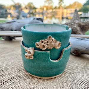 May include: Two-piece teal ceramic bowl set with coral-like embellishments. The top bowl has a rounded shape, while the bottom bowl has a cutout for chopsticks. The bowls are stacked, and chopsticks rest on top. The set is on a burlap surface.