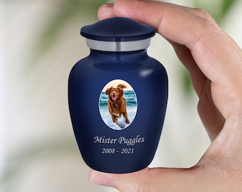 May include: A blue ceramic pet urn with a silver lid. The urn has a photo of a brown dog running on a beach inside a white oval. The urn is engraved with the text "Mister Puggles 2008 - 2021".
