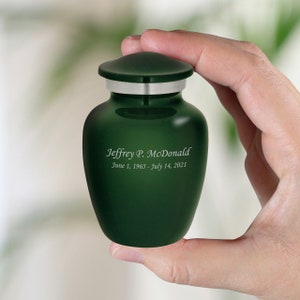 May include: A small, dark green urn with a silver lid. The urn is engraved with the name "Jeffrey P. McDonald" and the dates "June 1, 1965 - July 14, 2021".