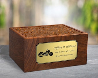 Könnte beinhalten: Eine rechteckige Urne aus Holz mit dunkelbraunem Finish und einem geschnitzten Blumenmuster auf dem Deckel. Eine goldfarbene Metallplatte auf der Vorderseite zeigt eine Motorradgrafik, den Namen Jeffrey P. Williams, Daten und Text.