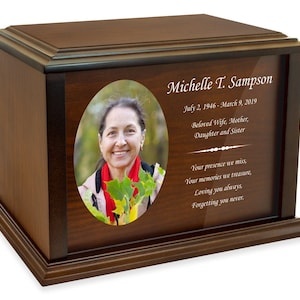 May include: A wooden cremation urn with a photo of a woman and an inscription that reads "Michelle T. Sampson July 2, 1946 - March 9, 2019 Beloved Wife, Mother, Daughter and Sister Your presence we miss, Your memories we treasure, Loving you always, Forgetting you never."
