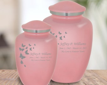 May include: Two pink cremation urns with silver lids. The urns are decorated with a butterfly design and engraved with the name "Jeffrey P. Williams" and dates. The text "My Love is Forever Yours" is also engraved on the urns.