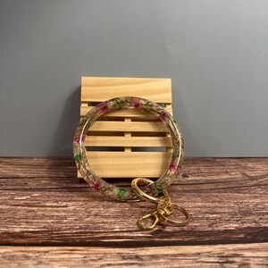 Pode incluir: Uma pulseira de chaveiro de resina transparente com flores secas embutidas em tons de rosa e verde. A pulseira é presa a um fecho e argolas de metal dourado. O fundo inclui um palete de madeira e uma superfície de madeira.