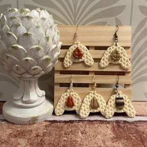 May include: Five dangle earrings shaped like cream-coloured cable knit sweaters, each featuring a unique central charm including two different leaves, two different pumpkins, and a coffee cup, all suspended from metal earring hooks on a wooden display.