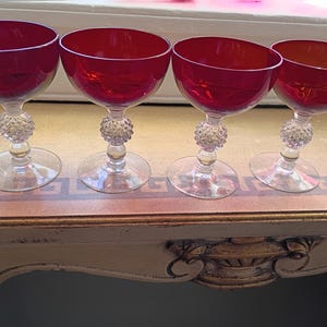 May include: Four vintage red champagne coupe glasses. Each glass has a round, clear base, a clear stem with a decorative, textured ball, and a wide, red bowl. The glasses are arranged on a decorative table.