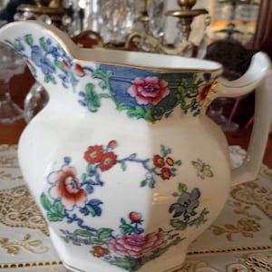 May include: White ceramic pitcher with a floral design featuring red, pink, and blue flowers, green leaves, and a blue border. The pitcher has a curved handle and a gold trim around the rim.