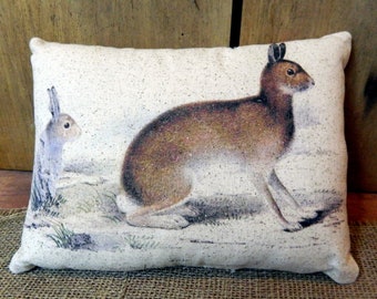 May include: Rectangular decorative pillow featuring a detailed illustration of two hares in a naturalistic setting. The larger hare is brown and tan, while the smaller one is gray. The pillow has a neutral background and a textured appearance, suitable for home decor.