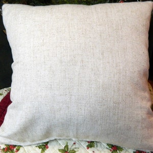 May include: A square, neutral-toned pillow with a textured, linen-like appearance. The pillow is a light beige color and appears soft. The pillow is placed on a patterned surface with red and green accents, suggesting a holiday theme.