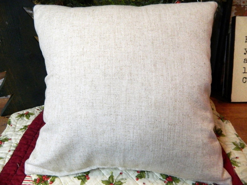 May include: A square, neutral-toned pillow with a textured, linen-like appearance. The pillow is a light beige color and appears soft. The pillow is placed on a patterned surface with red and green accents, suggesting a holiday theme.