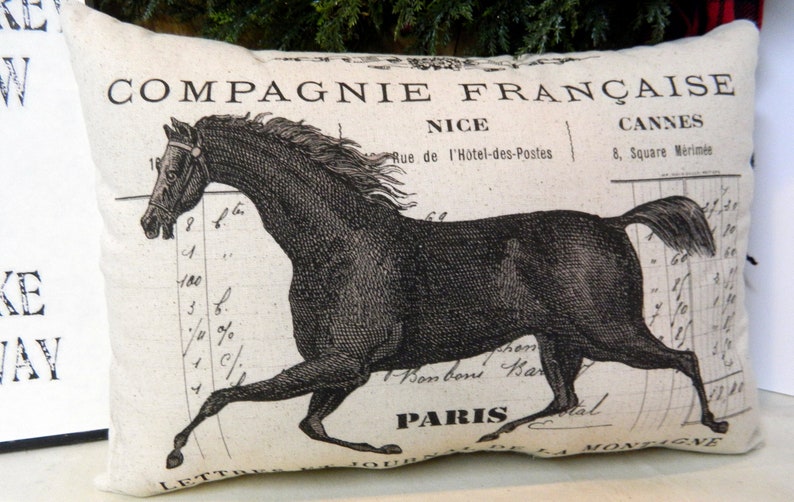 May include: Rectangular pillow with a black horse illustration. The pillow features French text, including "COMPAGNIE FRAN&Ccedil;AISE," "NICE," "CANNES," and "PARIS." The design has a vintage, antique aesthetic, with a cream-colored background.