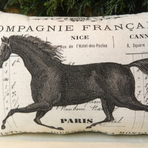 May include: Rectangular pillow with a vintage design featuring a black horse illustration. The pillow has French text, including "COMPAGNIE FRAN&Ccedil;AISE" and place names like "NICE" and "PARIS". The background is a cream color.