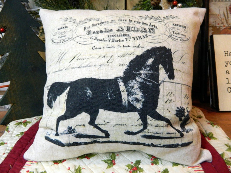 May include: A decorative pillow with a black horse silhouette and vintage French text. The pillow is beige and has a rectangular shape. The horse design is centered on the pillow, with the text above and below the horse.
