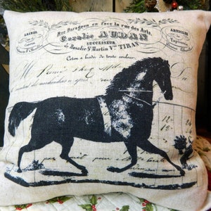 May include: A square decorative pillow with a black horse silhouette on a cream-colored background. The pillow features faded text and vintage-style graphics, including the name "Coralie AUDAN". The pillow is likely made of linen or a similar fabric.