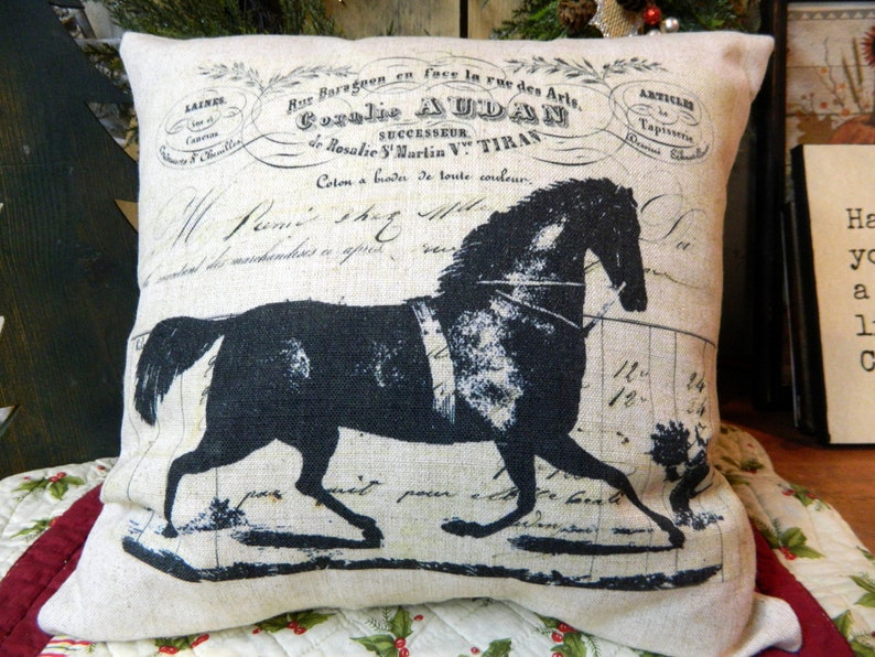 May include: A square decorative pillow with a black horse silhouette on a cream-colored background. The pillow features faded text and vintage-style graphics, including the name "Coralie AUDAN". The pillow is likely made of linen or a similar fabric.