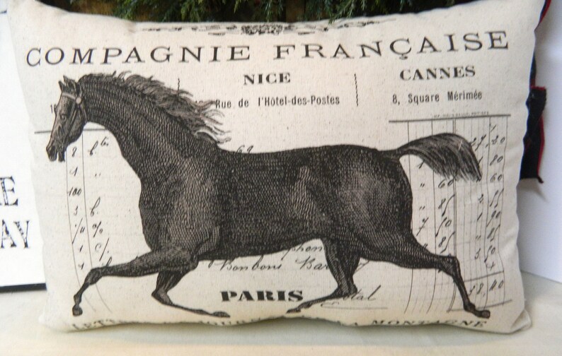 May include: Rectangular decorative pillow with a black and white illustration of a running horse. The pillow has French text, including "COMPAGNIE FRAN&Ccedil;AISE" and the city names NICE and CANNES. The design has a vintage, antique aesthetic.