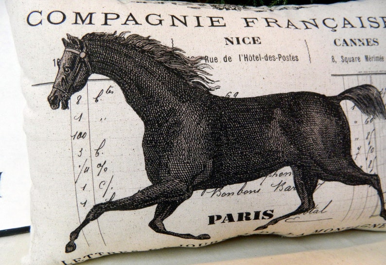 May include: Cream-colored decorative pillow featuring a black illustration of a running horse. The pillow has French text and numbers printed on it, with the words "COMPAGNIE FRAN&Ccedil;AISE", "NICE", "CANNES", and "PARIS" visible.