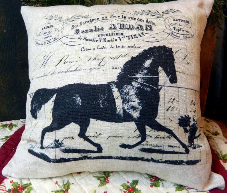 May include: A decorative pillow with a black horse silhouette and vintage French script. The pillow has a neutral linen-like background with printed text and a horse design, suitable for home decor.