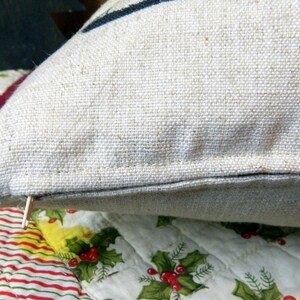 May include: Close-up of a beige linen pillow with a dark blue stripe. The pillow has a zipper closure and is resting on a quilted surface with a Christmas-themed pattern of holly and berries.