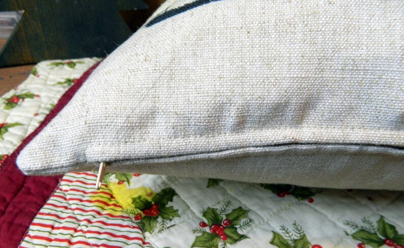 May include: Close-up of a beige linen pillow with a dark blue stripe. The pillow has a zipper closure and is resting on a quilted surface with a Christmas-themed pattern of holly and berries.