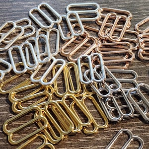 Puede incluir: Primer plano de una colección de deslizadores de tirantes de sujetador metálicos en tonos oro, plata y oro rosa. Los deslizadores rectangulares están dispuestos en primer plano, mostrando sus superficies lisas y brillantes. Estos accesorios se utilizan en lencería y confección de prendas.