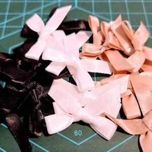 May include: A collection of small satin bows in various colors, including white, black, and a soft peach. The bows are neatly tied and arranged, suggesting their use in crafts or embellishments. The number 60 is visible on the cutting mat.