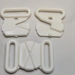 Puede incluir: Tres piezas de plástico blanco para la sujeción de tirantes de sujetador. Dos piezas tienen un lazo rectangular y un diseño en forma de gancho. La tercera pieza tiene dos lazos y una sección central con un diseño geométrico.