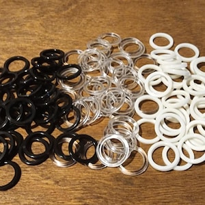 May include: An assortment of small, circular rings in three colors: black, clear, and white. The rings are scattered on a wooden surface, with the black rings clustered on the left, clear in the center, and white on the right.