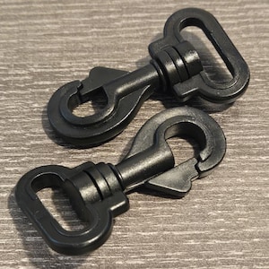 May include: Two black swivel snap hooks. Each hook has a spring-loaded gate and a rectangular loop for attachment. The hooks are made of a durable plastic material and are designed for various applications.