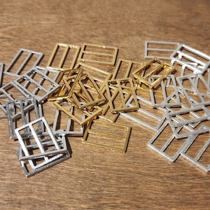 May include: A collection of rectangular metal jewelry findings in gold and silver tones. The findings are arranged on a wooden surface, with a variety of open frame designs. The pieces are small and appear to be components for jewelry making or crafting projects.