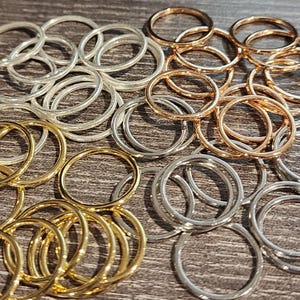 以下が含まれることがあります： 金、銀、ローズゴールドの色合いの金属リングのコレクション。リングはシンプルな円形デザインで、木製の表面に散らばっています。照明はジュエリーの金属的な輝きを強調しています。