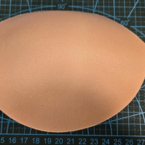 Puede incluir: Una copa de sujetador de color melocotón claro, de forma ovalada, sobre una esterilla de corte negra con líneas de cuadrícula y marcas de grados. La copa del sujetador es lisa y parece estar hecha de espuma o material similar.