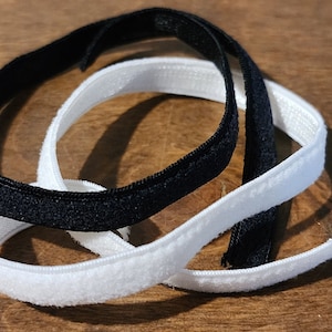 May include: Close-up of two elastic bands, one black and one white, lying on a wooden surface. The bands are thin and flexible, with a soft, velvety texture. The black band is slightly thicker than the white band.