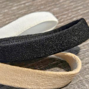 May include: Close-up of three elastic headbands in white, black, and beige. The black headband has a textured, velvety appearance. The white headband is smooth and the beige one has a woven texture. The headbands are slightly curved.