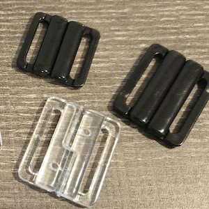 May include: Close-up of three bra strap adjusters. Two are black, and one is clear. The adjusters have a rectangular shape with a central bar and two loops on either side. They are made of plastic and are designed to adjust the length of bra straps.