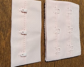 May include: Two pale pink bra extenders with white hook and eye closures. One extender has three hooks, the other has three eyes. The extenders are made of a soft, textured fabric and are designed to add extra length to a bra band.