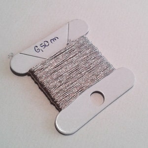 May include: A spool of silver metallic thread, with the text "6,50 m" written on the silver plastic spool. The thread is tightly wound and appears to be made of many fine strands. The spool has a hole for hanging.