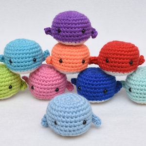 May include: A collection of nine crocheted whale plushies in various colors, including blue, green, pink, red, orange, and purple. Each whale has black eyes and a white belly.
