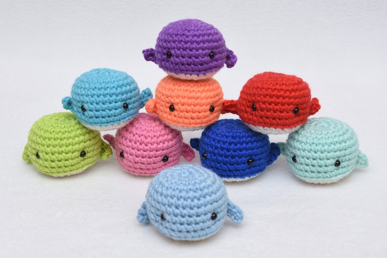 May include: A collection of nine crocheted whale plushies in various colors, including blue, green, pink, red, orange, and purple. Each whale has black eyes and a white belly.