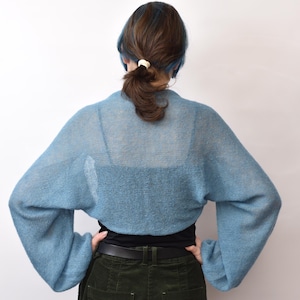 May include: A light blue, sheer, knitted bolero with long, loose sleeves and a cropped length. The garment features a fine, open-knit texture that allows the underlying black top to be visible through the fabric, creating a layered appearance.
