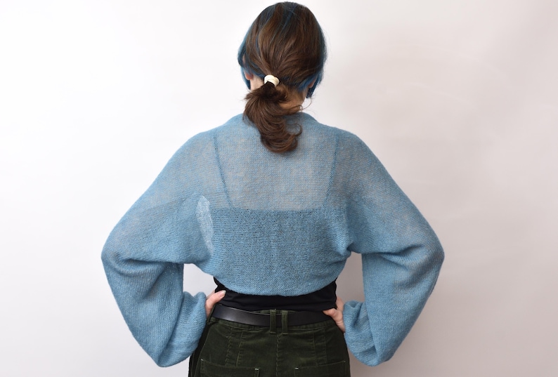 May include: A light blue, sheer, knitted bolero with long, loose sleeves and a cropped length. The garment features a fine, open-knit texture that allows the underlying black top to be visible through the fabric, creating a layered appearance.