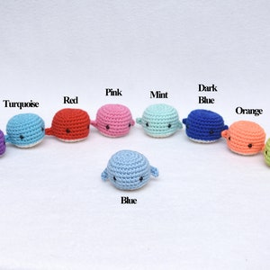 May include: A set of ten crocheted whale toys in various colors, including purple, turquoise, red, pink, mint, dark blue, orange, and green. The whales are arranged in a semi-circle with a light blue whale in the center.