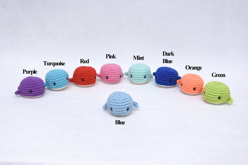 May include: A set of ten crocheted whale toys in various colors, including purple, turquoise, red, pink, mint, dark blue, orange, and green. The whales are arranged in a semi-circle with a light blue whale in the center.