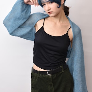 May include: A light blue, long-sleeved, open-front knit bolero shrug made of a soft, sheer material. The garment features voluminous sleeves with ribbed cuffs, worn over a black camisole and dark green trousers with a black leather belt.