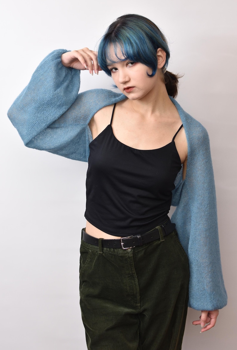 May include: A light blue, long-sleeved, open-front knit bolero shrug made of a soft, sheer material. The garment features voluminous sleeves with ribbed cuffs, worn over a black camisole and dark green trousers with a black leather belt.
