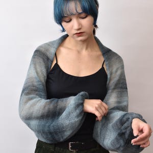 May include: A soft, knitted shrug featuring a gradient of blue, gray, and black tones. The garment has long, voluminous sleeves with gathered cuffs and an open-front design that drapes across the shoulders and arms, showcasing a textured knit.