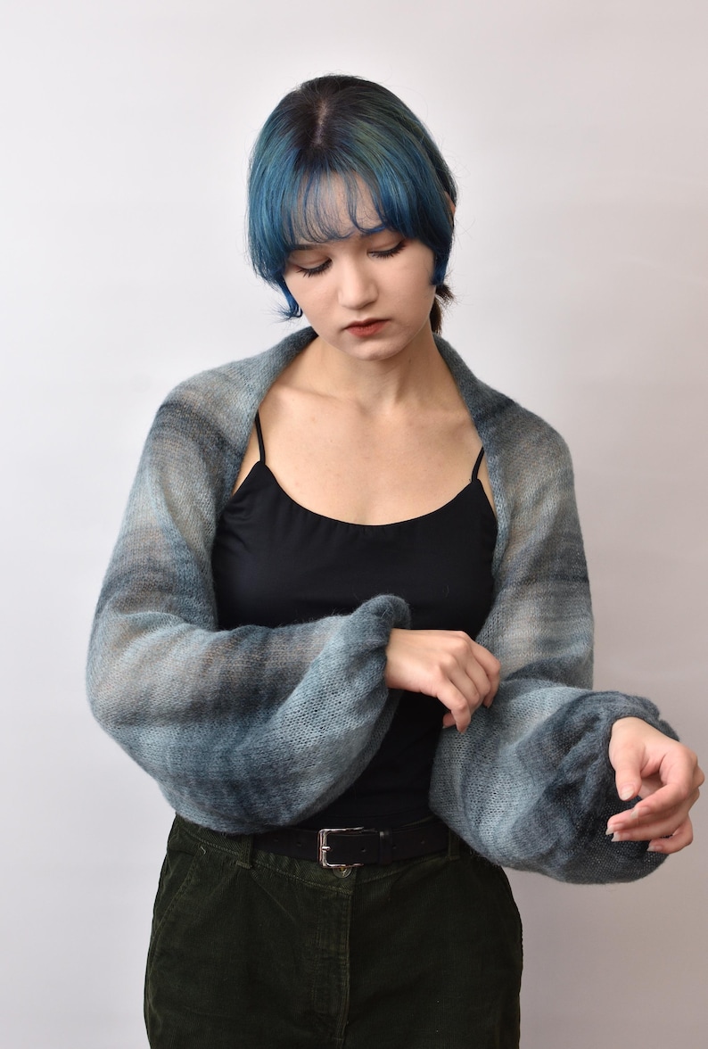 May include: A soft, knitted shrug featuring a gradient of blue, gray, and black tones. The garment has long, voluminous sleeves with gathered cuffs and an open-front design that drapes across the shoulders and arms, showcasing a textured knit.