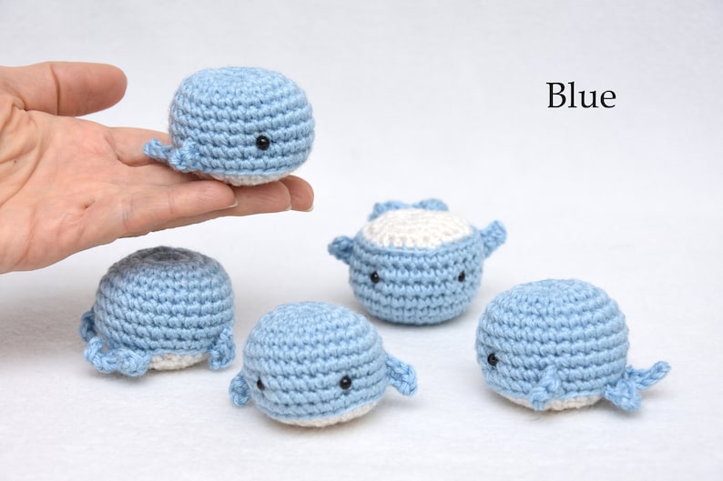 May include: Five crocheted whale plushies in blue and white. The whales have black eyes and are arranged on a white surface. Blue.