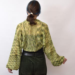 May include: A cropped, long-sleeved knit bolero in a variegated lime green and dark olive pattern. The garment features a loose, open-knit texture with wide sleeves and a rounded back hem, worn over a solid black top and dark green corduroy pants.
