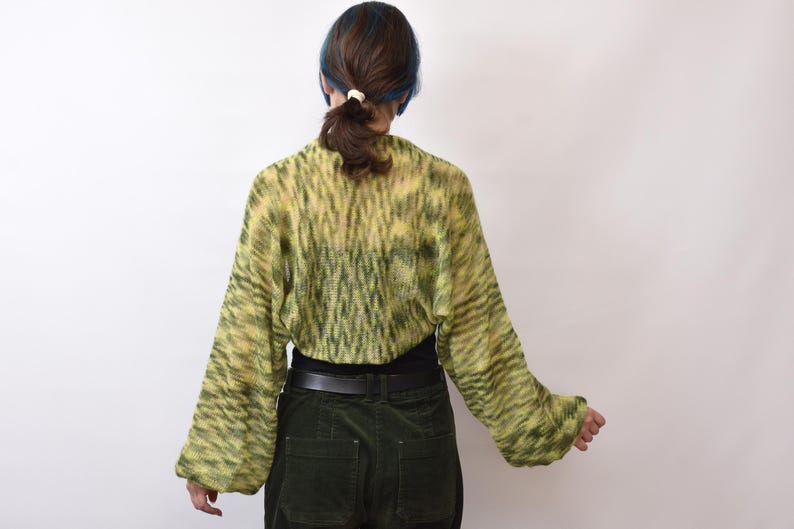 May include: A cropped, long-sleeved knit bolero in a variegated lime green and dark olive pattern. The garment features a loose, open-knit texture with wide sleeves and a rounded back hem, worn over a solid black top and dark green corduroy pants.