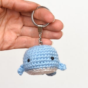 May include: A light blue crocheted whale keychain with black eyes. The whale has a white belly and is attached to a silver key ring.